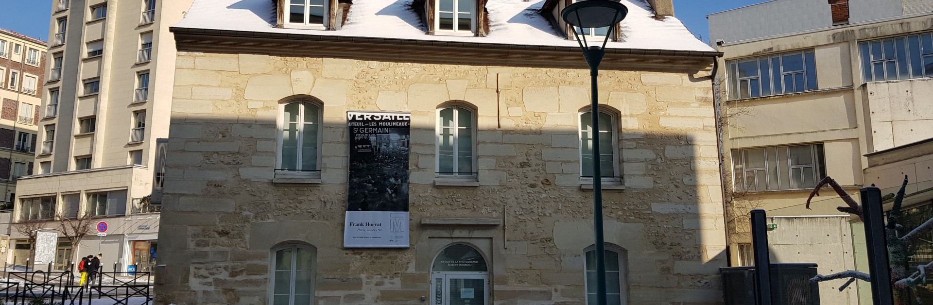 Une maison en pierre beige au toit recouvert de neige, avec une affiche devant et l